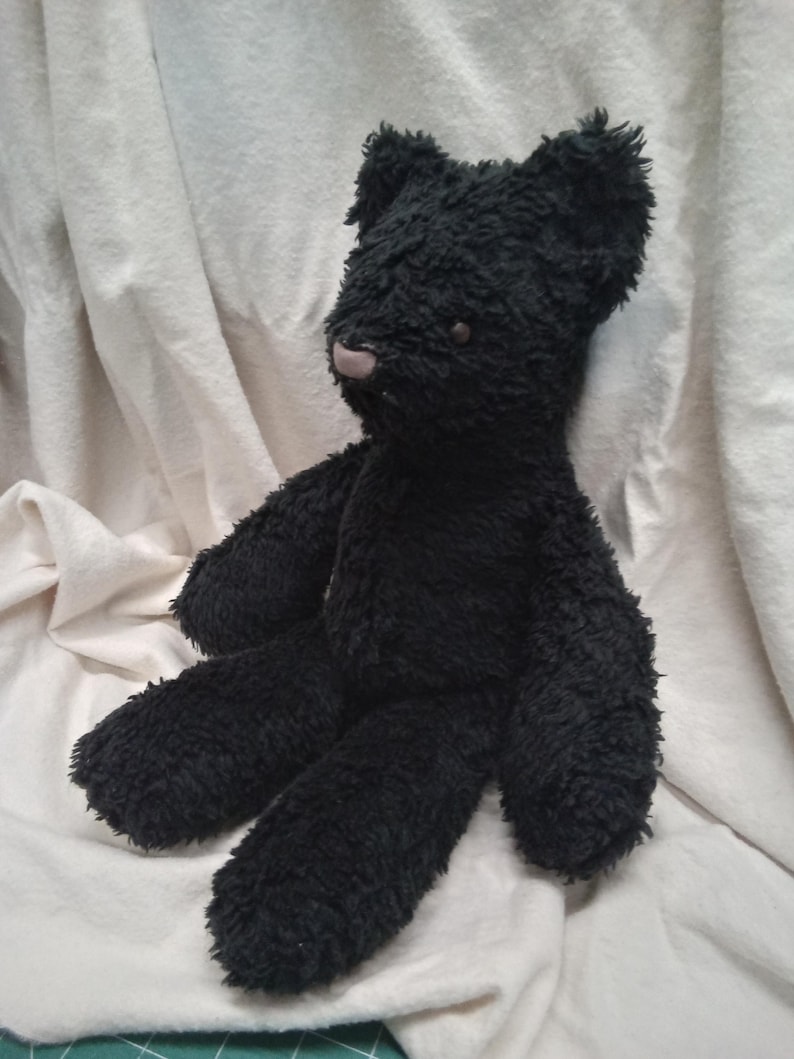 May include: A handmade black teddy bear with a soft, textured appearance. The bear has a light pink nose and small, round eyes. Its arms and legs are extended, and it is sitting on a white surface.