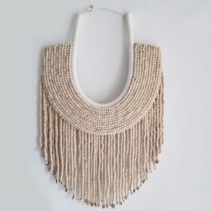 May include: A beige beaded necklace with a white fabric backing. The necklace features a curved upper section and long, dangling strands of beads. The beads are a light, natural color, and the necklace hangs against a white background.
