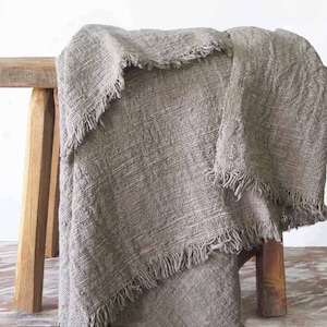 May include: A grey linen throw blanket with a fringed edge draped over a wooden stool.