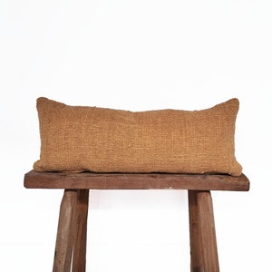 May include: A long, rectangular, brown throw pillow with a woven texture sits on a wooden bench.