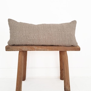 Puede incluir: Un cojín lumbar de lino marrón claro descansando sobre un banco de madera con un acabado rústico.