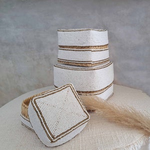 May include: Three white beaded boxes with gold trim, stacked on top of each other. The boxes are on a white surface with a dried grass stalk.