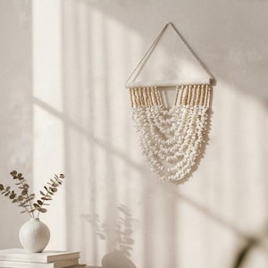 Puede incluir: Escena interior de tonos neutros con una consola de madera, una cesta tejida y un pequeño jarrón blanco con vegetación. También hay un tapiz de macramé y libros. La luz suave y las sombras crean un ambiente cálido y acogedor.