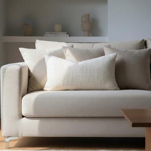 Könnte beinhalten: Ein cremefarbenes Sofa mit mehreren dekorativen Kissen in verschiedenen Beige- und Cremetönen. Das Sofa hat ein schlichtes, modernes Design mit einem niedrigen Profil und einem Holztisch davor. Der Raum ist hell und wirkt beruhigend.