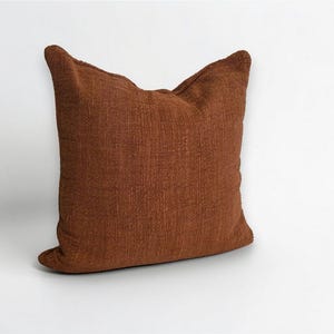 Puede incluir: Un coj&iacute;n cuadrado de color marr&oacute;n rojizo c&aacute;lido. El coj&iacute;n tiene una superficie texturizada y un aspecto suave, perfecto para a&ntilde;adir comodidad y estilo a cualquier espacio habitable. Es un art&iacute;culo decorativo.