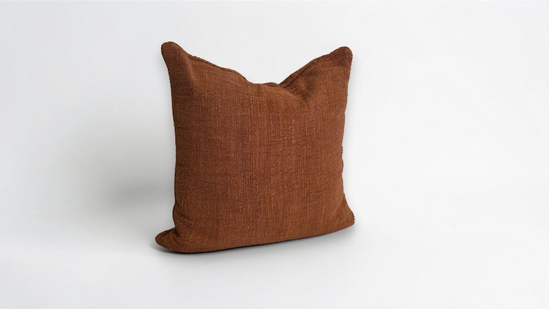 Puede incluir: Un coj&iacute;n cuadrado de color marr&oacute;n rojizo c&aacute;lido. El coj&iacute;n tiene una superficie texturizada y un aspecto suave, perfecto para a&ntilde;adir comodidad y estilo a cualquier espacio habitable. Es un art&iacute;culo decorativo.