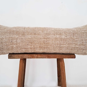 May include: A long, rectangular beige pillow with a subtle woven pattern. The pillow is sitting on a wooden stool.