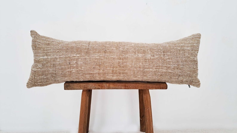 May include: A long, rectangular beige pillow with a subtle woven pattern. The pillow is sitting on a wooden stool.