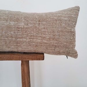 May include: A long, rectangular pillow with a beige and white woven fabric cover. The pillow is sitting on a wooden stool.