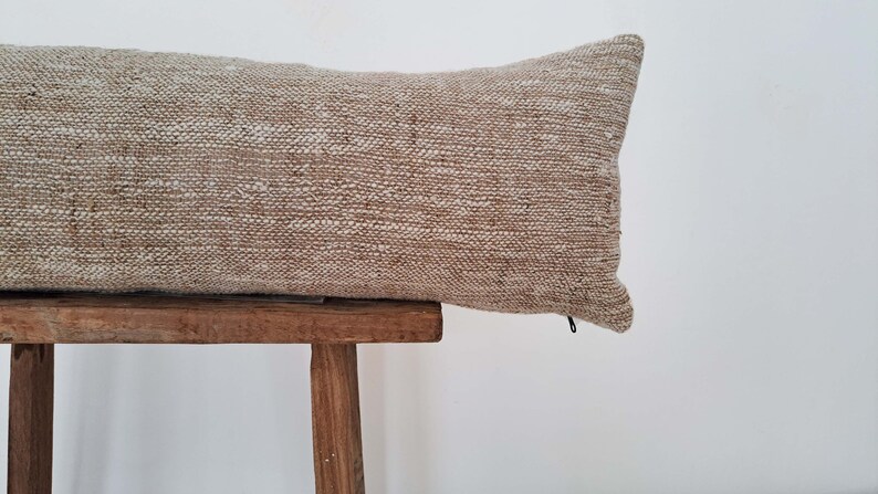 May include: A long, rectangular pillow with a beige and white woven fabric cover. The pillow is sitting on a wooden stool.