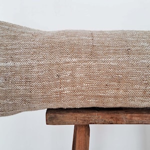 May include: A long, rectangular, beige pillow with a woven texture sits on a wooden stool. The pillow is made of a natural fiber, possibly linen or cotton.