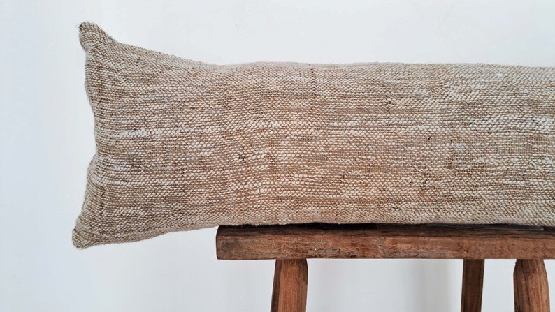 May include: A long, rectangular, beige pillow with a woven texture sits on a wooden stool. The pillow is made of a natural fiber, possibly linen or cotton.