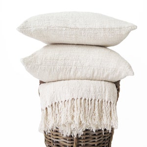 May include: A stack of cream-colored pillows and a fringed throw blanket rest inside a woven basket. The pillows have a textured appearance, and the blanket's fringe hangs over the basket's edge. The basket is made of brown wicker.