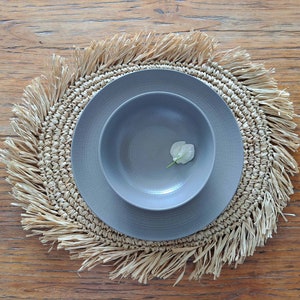 Pode incluir: Um conjunto de prato e tigela de cerâmica cinza em um descanso de mesa tecido natural com borda franjada. O descanso de mesa é redondo e de cor marrom natural.