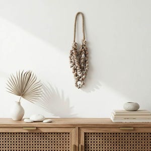 Puede incluir: Un colgante de pared decorativo hecho de conchas marinas, suspendido por una cuerda trenzada. Las conchas son de varios tonos beige y marrón. La imagen también muestra un mueble de madera con frente tejido, un jarrón con una hoja de palma seca y una pila de libros.