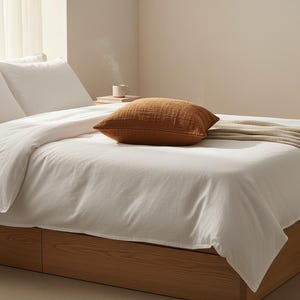 Puede incluir: Una cama con ropa de cama blanca, dos almohadas blancas y una almohada marr&oacute;n texturizada. Una manta beige est&aacute; sobre la cama. El marco de la cama es de madera. Una pila de libros y una taza est&aacute;n en el suelo.