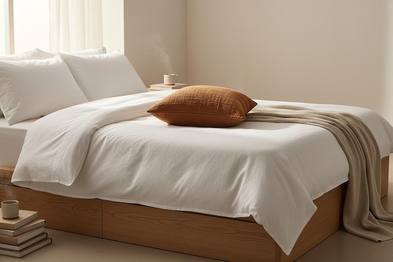 Puede incluir: Una cama con ropa de cama blanca, dos almohadas blancas y una almohada marr&oacute;n texturizada. Una manta beige est&aacute; sobre la cama. El marco de la cama es de madera. Una pila de libros y una taza est&aacute;n en el suelo.