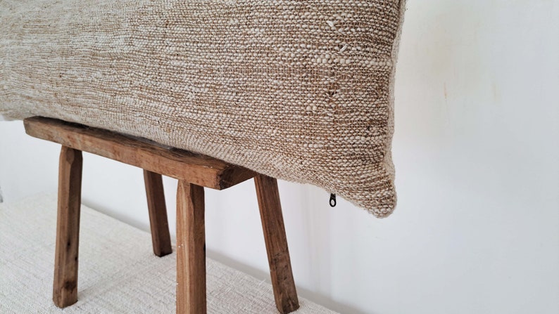 May include: A beige rectangular pillow with a zipper closure on a wooden stool. The pillow is made of a textured fabric with a subtle sheen.