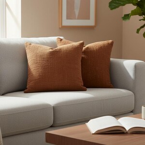 Puede incluir: Un sof&aacute; gris claro con dos cojines decorativos texturizados de color &oacute;xido. Una manta color crema est&aacute; sobre el brazo. Una mesa de centro de madera tiene un libro abierto y una taza. Una planta en maceta est&aacute; en el fondo.