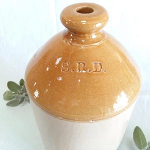 Puede incluir: Una jarra de gres vintage en tonos beige y crema. La jarra tiene un cuerpo redondeado, un cuello estrecho y una pequeña abertura en la parte superior. Las letras "S.R.D." están grabadas en la parte delantera. La jarra se utiliza probablemente para almacenar líquidos.