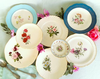 May include: A collection of vintage floral plates in various sizes and designs. The plates feature colorful floral patterns, including roses and daisies, with gold trim. Some plates have a blue or white scalloped edge. The arrangement includes pink flowers and greenery.