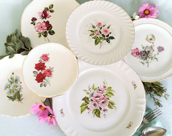 May include: Five white ceramic plates with gold trim and floral designs. The plates feature pink, red, and purple flowers with green leaves. The plates are arranged in a circle on a light blue surface.