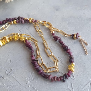 Op de afbeelding: Een gelaagde ketting met goudkleurige schakels en strengen kleine, donkerpaarse edelstenen. Goudkleurige, onregelmatig gevormde kralen zijn door het ontwerp verspreid. De ketting ligt op een lichtgrijze ondergrond.