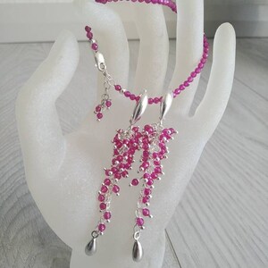 May include: A set of fuchsia-coloured jewellery, including a necklace, earrings, and bracelet. The necklace and bracelet are made of small, round beads. The earrings feature cascading clusters of beads with teardrop-shaped silver accents. The jewellery is displayed on a white hand.