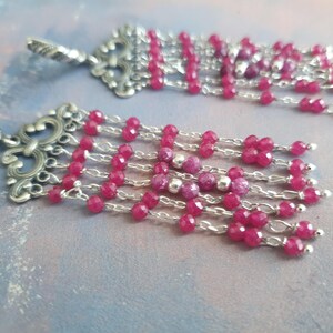 Op de afbeelding: Een paar zilverkleurige oorhangers met een filigraan ontwerp. Elke oorbel heeft meerdere strengen kleine, helder roze kralen, afgewisseld met zilveren kralen, waardoor een watervaleffect ontstaat. De oorbellen worden gepresenteerd op een getextureerd oppervlak.