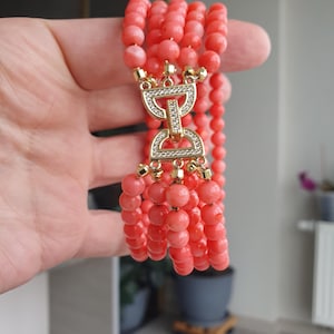 May include: A coral-colored beaded bracelet with a gold-colored clasp. The bracelet features multiple strands of round beads. The clasp is adorned with clear crystals. The jewelry is held up against a neutral background.