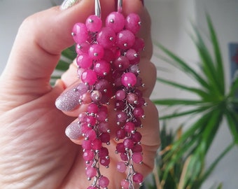 May include: A pair of long, dangling earrings featuring clusters of faceted, round, hot pink and magenta beads. The beads are attached to thin silver-toned metal chains, creating a cascading, multi-strand effect that tapers toward the bottom.