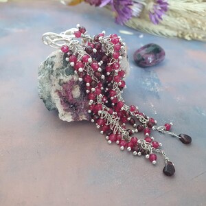 May include: Silver earrings with cascading strands of small, round, red beads. The earrings are draped over a textured, gray and pink stone. A small, polished, purple stone sits nearby, with purple flowers in the background.