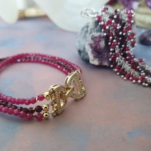 Peut inclure: Un bracelet à trois rangs avec un fermoir doré et de petites perles rondes rouges. Un pendentif assorti avec un groupe de perles rouges et des chaînes argentées est également visible. Les bijoux sont exposés sur une surface rose clair et bleue.