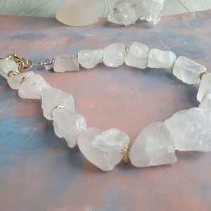Pode incluir: Uma pulseira de quartzo transparente com pedras de quartzo brutas e irregulares. As pedras são unidas com detalhes metálicos dourados e um fecho. A pulseira é exibida em uma superfície com tons de azul, rosa e branco.
