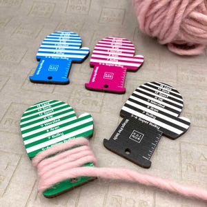 Könnte beinhalten: Drei hölzerne Strickmaschenzähler in Form von Fäustlingen. Jeder Maschenzähler hat eine andere Farbe: blau, rosa und schwarz. Die Maschenzähler haben einen Lineal an der Seite und ein gestreiftes Muster auf der Vorderseite. Die Maschenzähler sind mit verschiedenen Garnstärken beschriftet, wie z. B. Lace, Sock, Sport, DK, Worsted, Aran, Bulky und Super Bulky.
