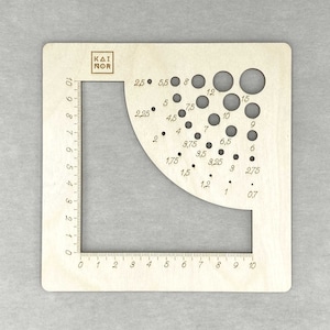 May include: A wooden ruler with a circular cutout featuring various sized holes marked with measurements in inches. The ruler is marked with numbers from 0 to 10 on both sides.