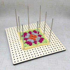Könnte beinhalten: Ein Holz-Blockbrett mit einem bunten gehäkelten Quadrat. Das Quadrat besteht aus rosa, orange, grau und grüner Wolle. Metallstifte sind um das Quadrat herum eingesetzt. Das Brett hat gleichmäßig verteilte Löcher. Der Text "KAI NOR" ist sichtbar.