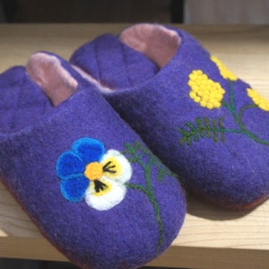 May include: A pair of purple felted slippers with a light pink interior lining. The left slipper features a blue, white, and yellow pansy flower design, while the right slipper displays a yellow mimosa flower branch with green stems.