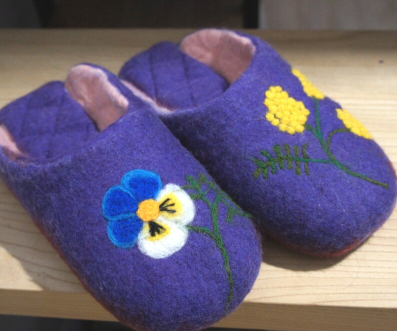 May include: A pair of purple felted slippers with a light pink interior lining. The left slipper features a blue, white, and yellow pansy flower design, while the right slipper displays a yellow mimosa flower branch with green stems.