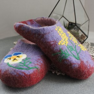 May include: A pair of felted wool slippers in shades of purple and deep red. The front slipper features a white and blue pansy flower embroidery, while the back slipper displays a yellow mimosa flower design with green stems and leaves.