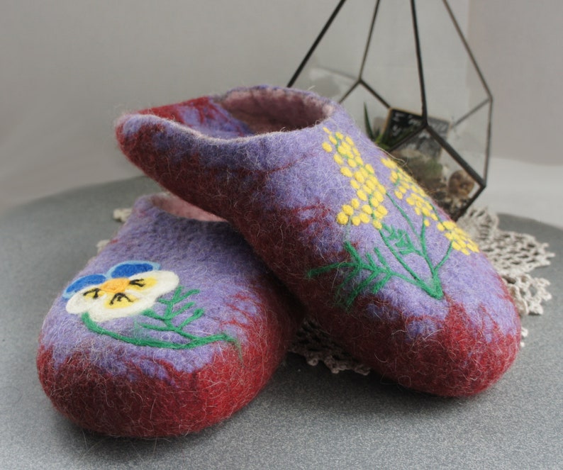 May include: A pair of felted wool slippers in shades of purple and deep red. The front slipper features a white and blue pansy flower embroidery, while the back slipper displays a yellow mimosa flower design with green stems and leaves.