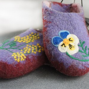May include: A pair of felted wool slippers in shades of purple and deep red. One slipper features an embroidered yellow mimosa branch, while the other displays a large, detailed pansy flower with blue, white, and yellow petals and green stems.
