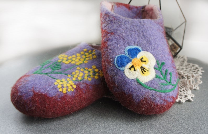 May include: A pair of felted wool slippers in shades of purple and deep red. One slipper features an embroidered yellow mimosa branch, while the other displays a large, detailed pansy flower with blue, white, and yellow petals and green stems.