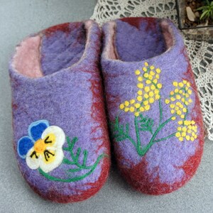 May include: A pair of felted wool slippers in shades of purple and deep red. The left slipper features a white, blue, and yellow pansy flower design, while the right slipper displays a yellow mimosa branch with green stems and leaves.