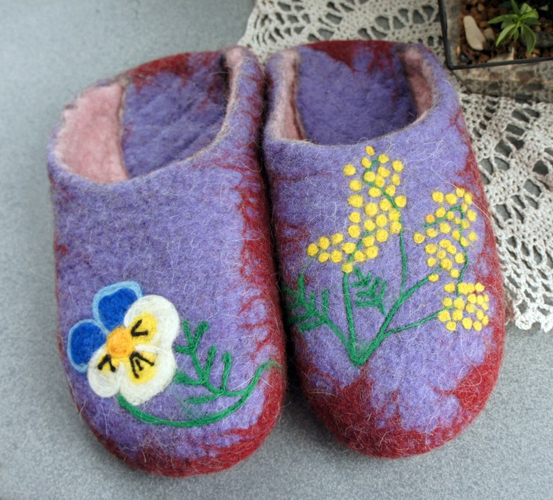 May include: A pair of felted wool slippers in shades of purple and deep red. The left slipper features a white, blue, and yellow pansy flower design, while the right slipper displays a yellow mimosa branch with green stems and leaves.