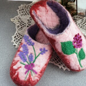 May include: A pair of felted wool slippers in shades of pink and deep red. The slippers feature a purple interior lining and a quilted footbed. One slipper displays a purple lavender sprig design, while the other shows a green leaf and purple flower.