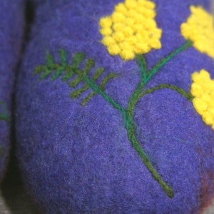 May include: A pair of purple felted wool slippers featuring an embroidered design of green stems and bright yellow mimosa flowers. The textured wool surface displays detailed needlework with vibrant yellow clusters and dark green foliage.