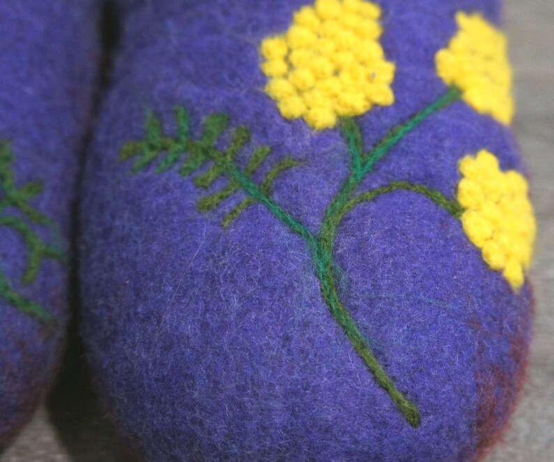 May include: A pair of purple felted wool slippers featuring an embroidered design of green stems and bright yellow mimosa flowers. The textured wool surface displays detailed needlework with vibrant yellow clusters and dark green foliage.