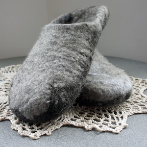 May include: A pair of grey felted wool slippers with a textured, fuzzy surface and a rounded toe design. The slippers feature a mottled grey color palette with darker charcoal tones near the base and a slip-on construction with an open heel.