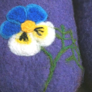 May include: A deep purple felted wool slipper featuring a needle-felted pansy flower design. The flower has bright blue upper petals, white lower petals with yellow centers, and dark markings. A thin green stem is stitched onto the purple surface.